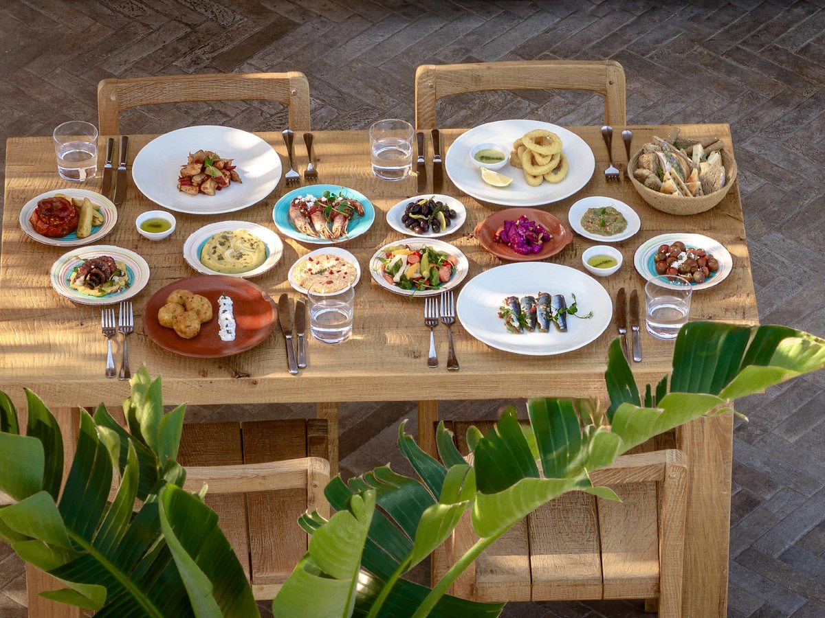 A wooden table laid with shared meze plates, dips, bread and seafood, set among tropical planting.