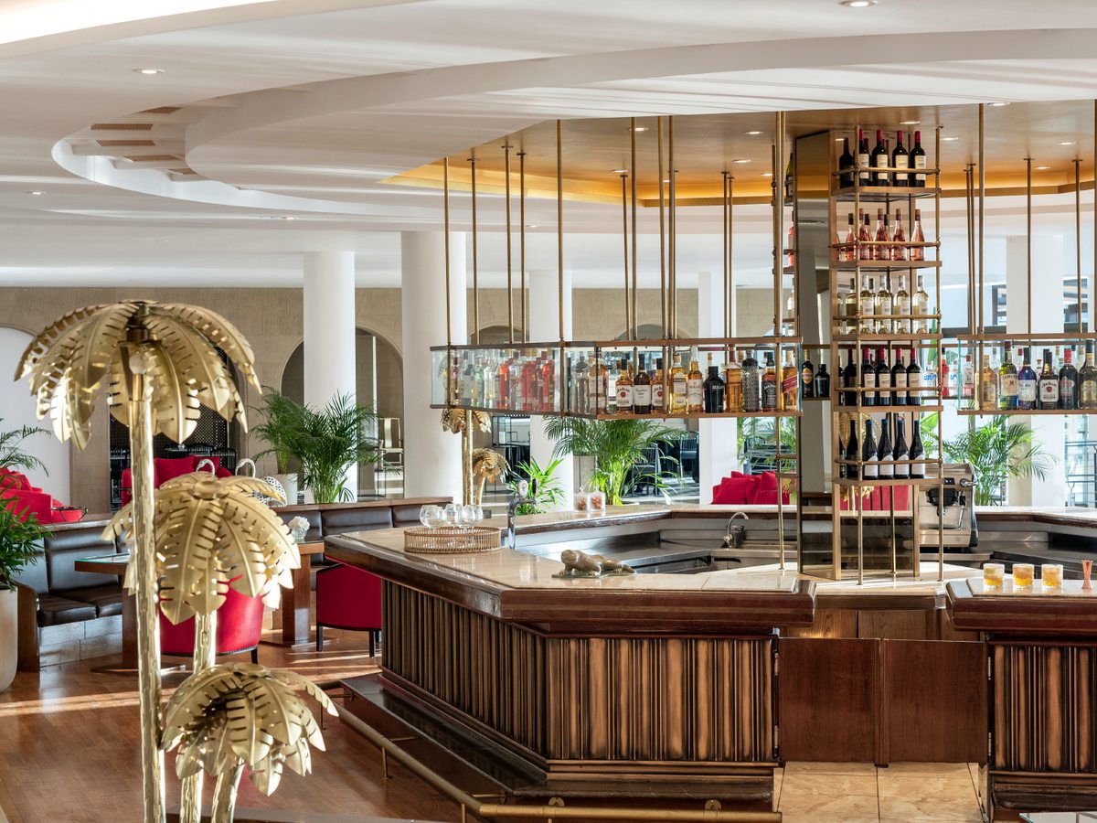 The circular lobby bar under a curved ceiling, with a suspended glass spirits gantry, brass palm-tree lamps and red lounge chairs.