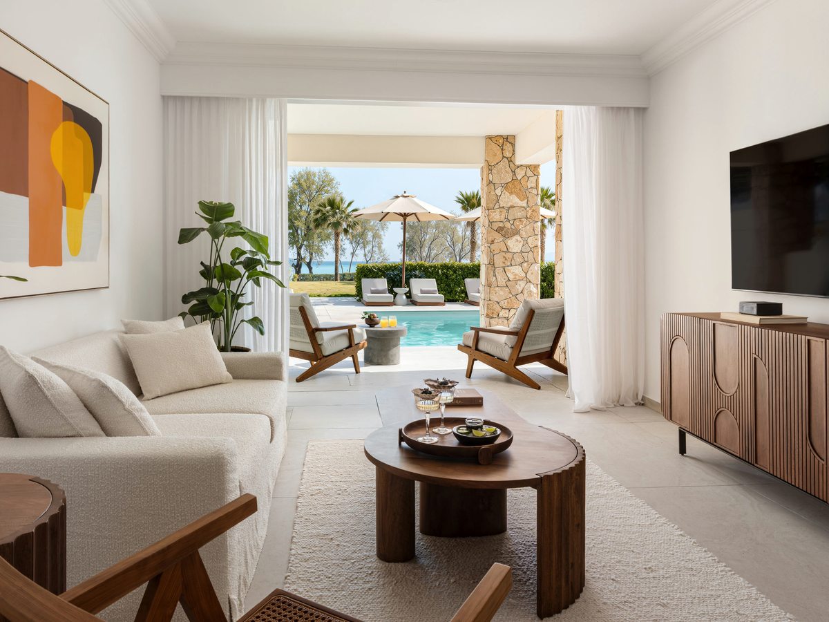 Suite living room with a cream sofa, wooden armchairs and a round timber coffee table on a pale rug, its doors folded back to a terrace with loungers, a private pool and the sea.