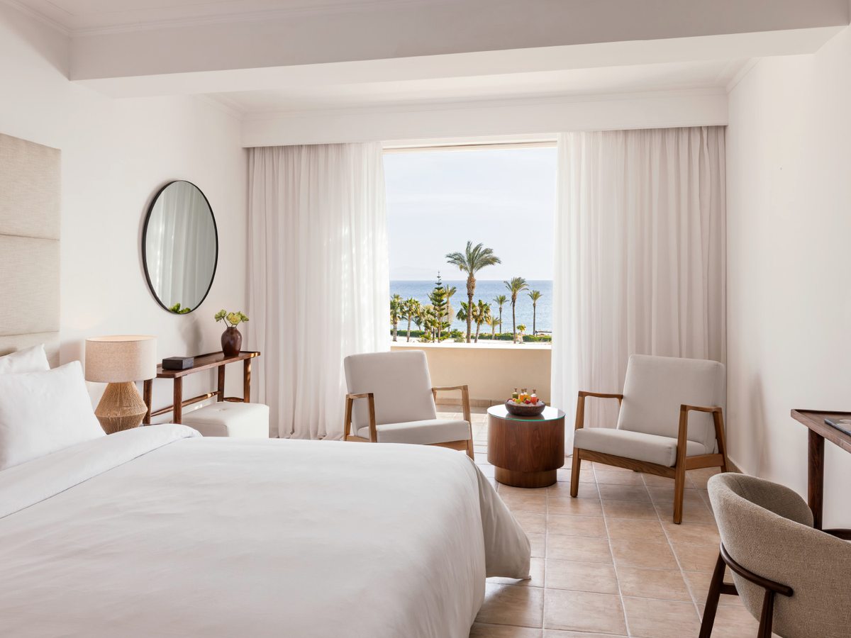 White-walled double room with a large bed, a round mirror, pale armchairs and a low table, its curtained doors open to a terrace with palm trees and the sea beyond.