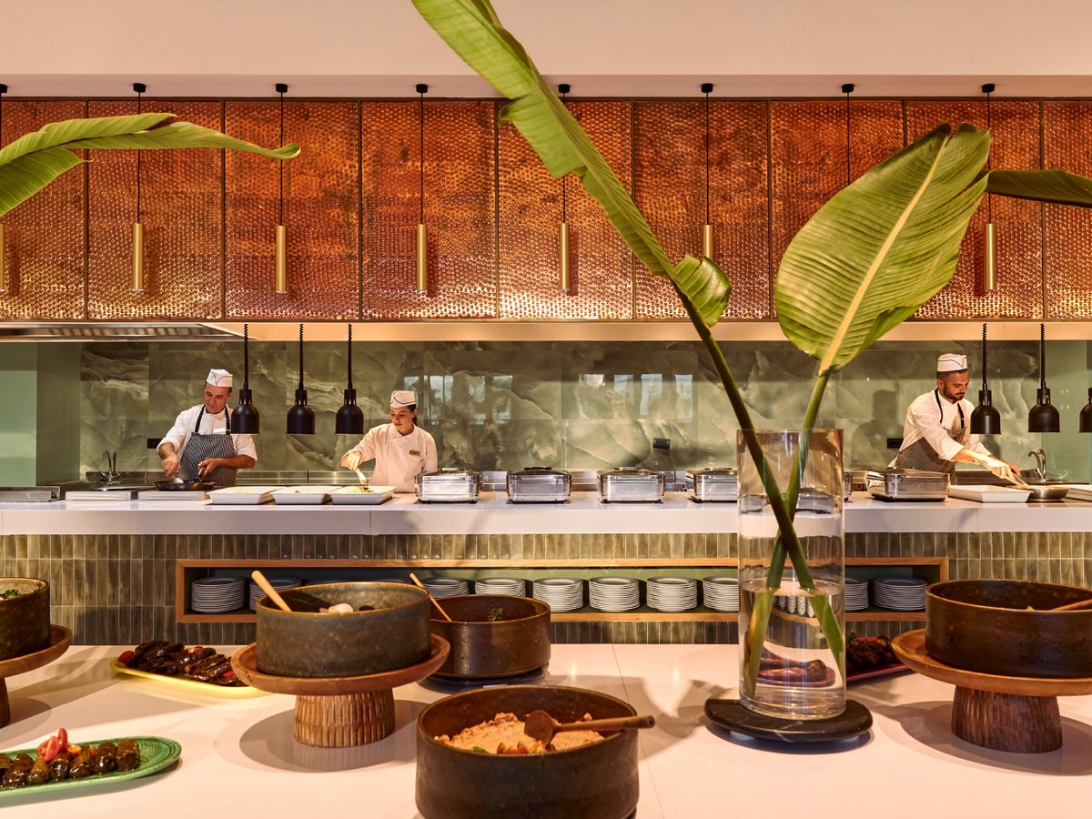 Buffet show-cooking counter with three chefs in whites plating dishes beneath copper-mesh cabinets and pendant heat lamps, clay serving bowls set out on a white counter in front.
