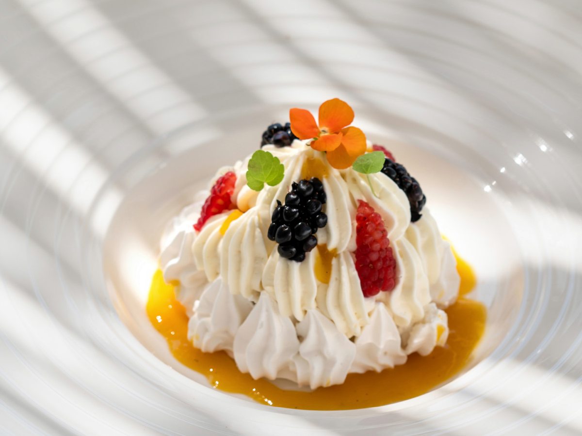 A domed meringue-and-cream dessert topped with blackberries, raspberries and a small orange flower, in a swirl of passion-fruit sauce on a white ridged plate.