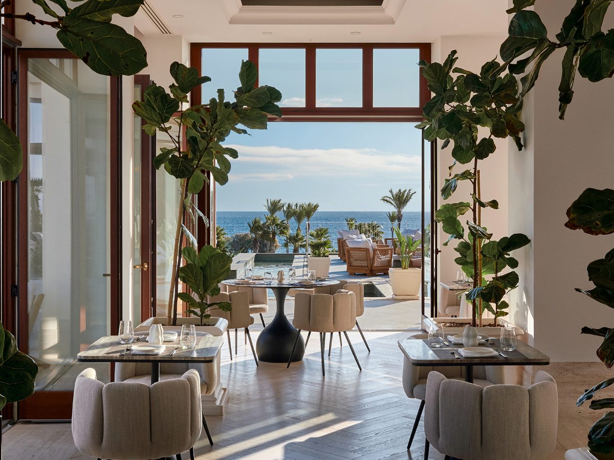 Bright restaurant dining room with pale tub chairs at marble tables around a round central table, framed by tall fiddle-leaf fig plants, its doors folded open to a sea-view terrace.