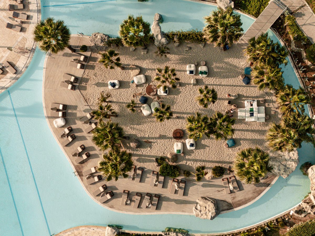 Aerial view of a sandy island dotted with palms, loungers and cabanas, encircled by the pale blue channel of a lazy river.