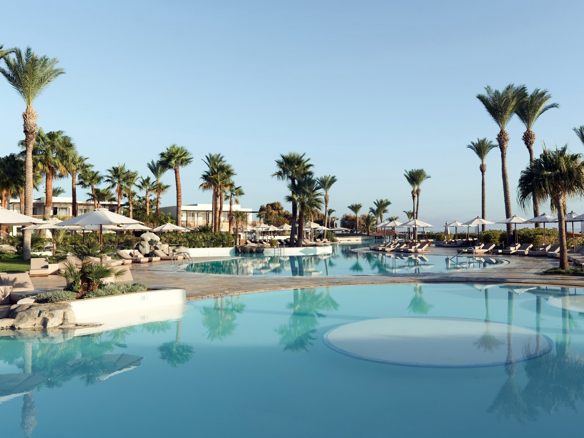 Wide lagoon-shaped freshwater pool with a shallow round platform in the foreground, ringed by palms, parasols and low white buildings under a clear sky.