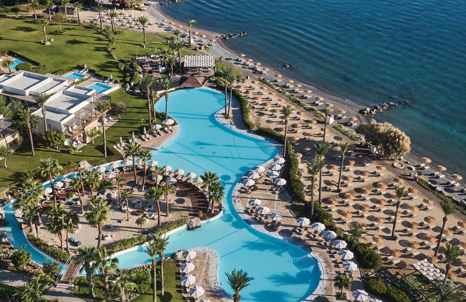 Grecotel LUXME Kos at Psalidi: the lagoon pool and its sandy island, ringed by palms and low bungalows, with the Aegean beyond.