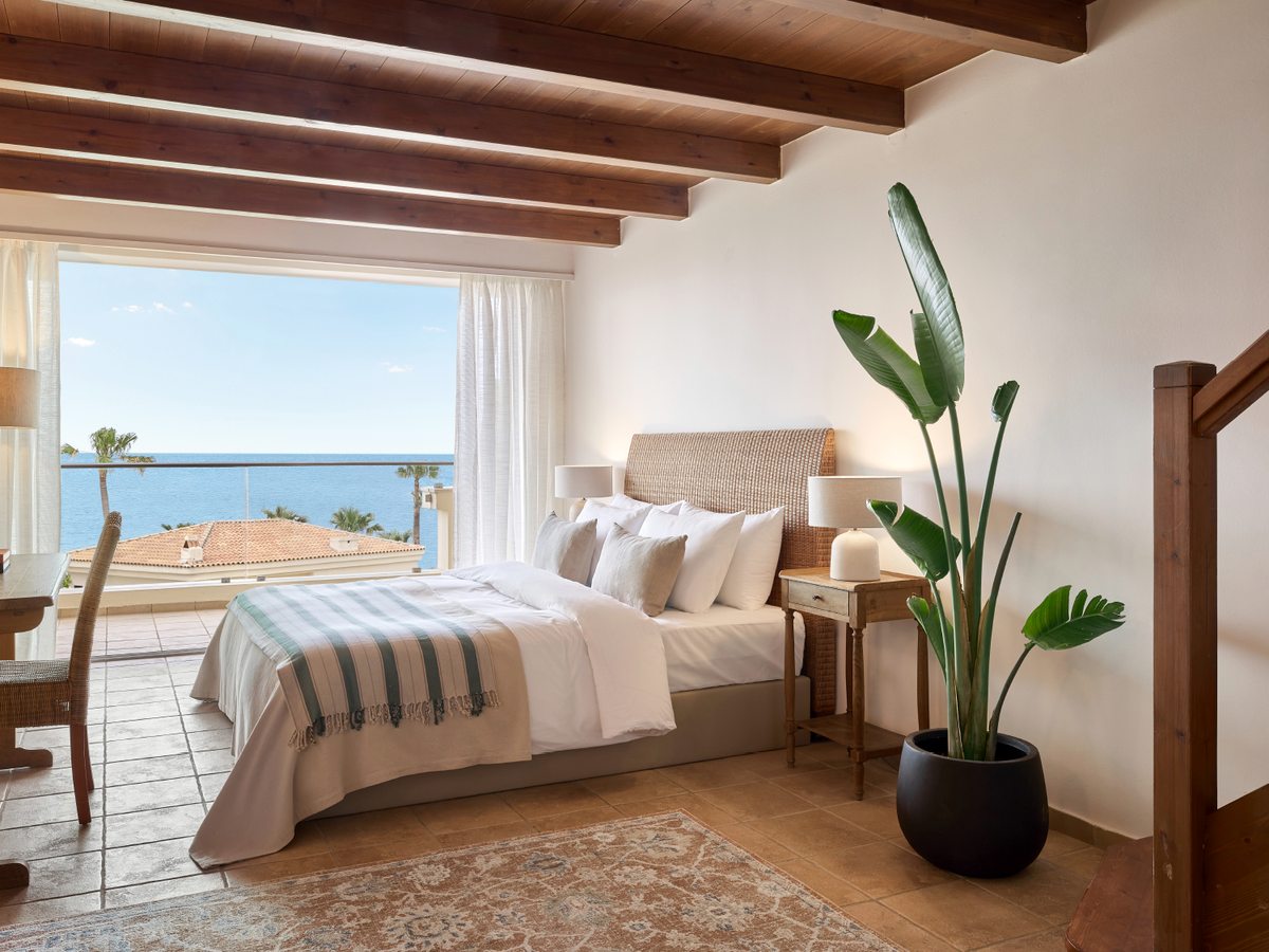 Bright bedroom with a timber-beamed ceiling and a large window framing the sea, a desk and a potted plant.