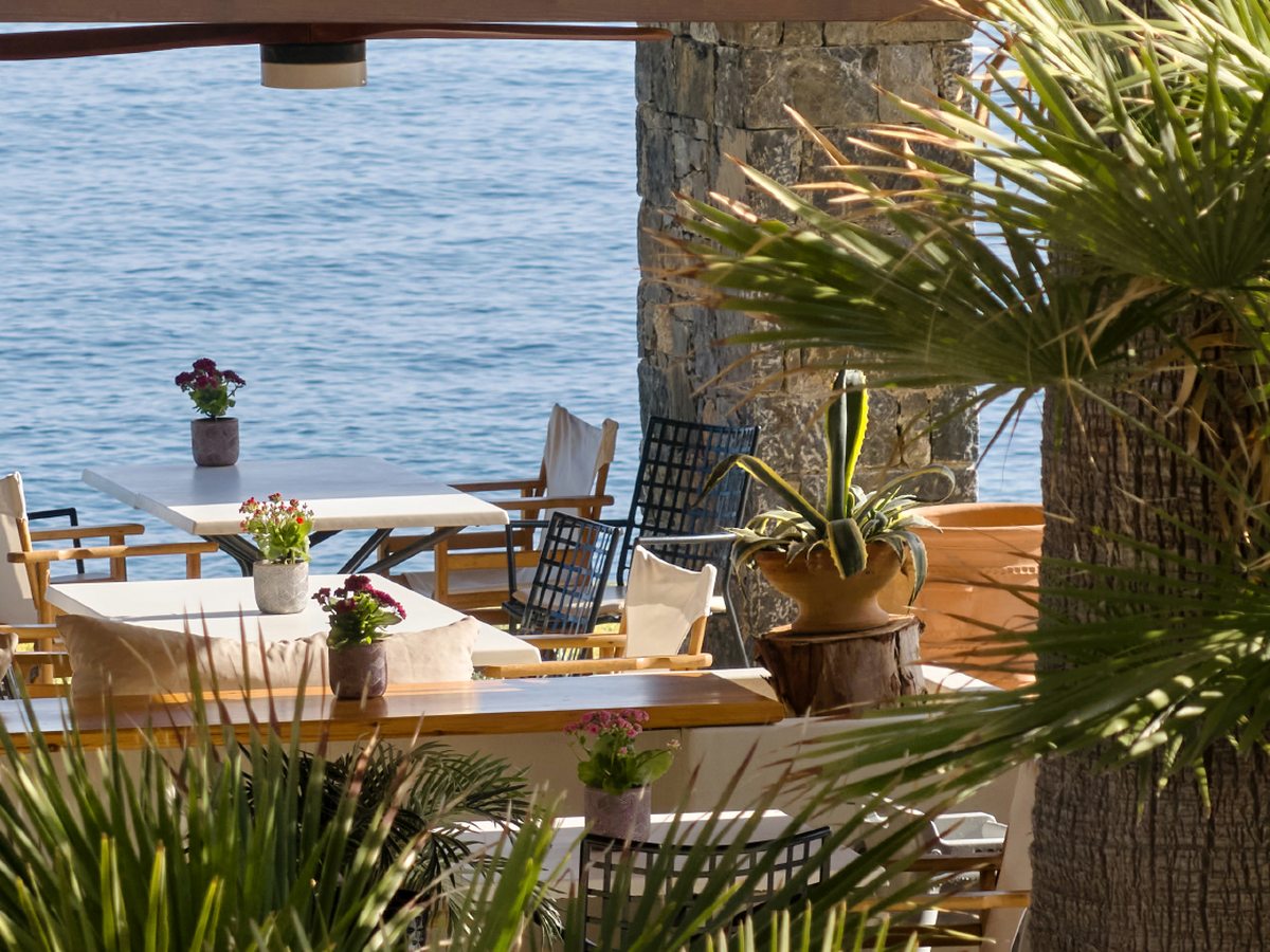 Open-air restaurant terrace with white-clothed tables under shade, looking out over the sea.