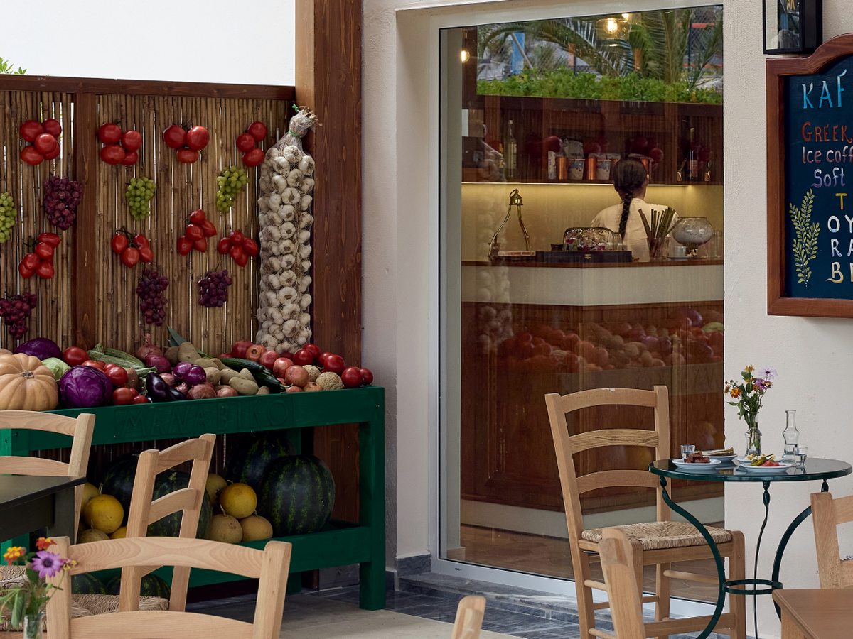 Outdoor cafe tables and chairs beside a rustic produce display and a chalkboard drinks menu at the Kafeneio coffee house.