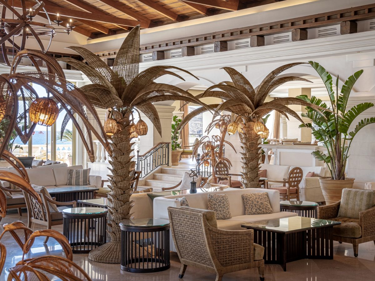 Spacious lobby lounge with sofas and armchairs among tall palm-tree columns under a timber ceiling, with large windows to the grounds.
