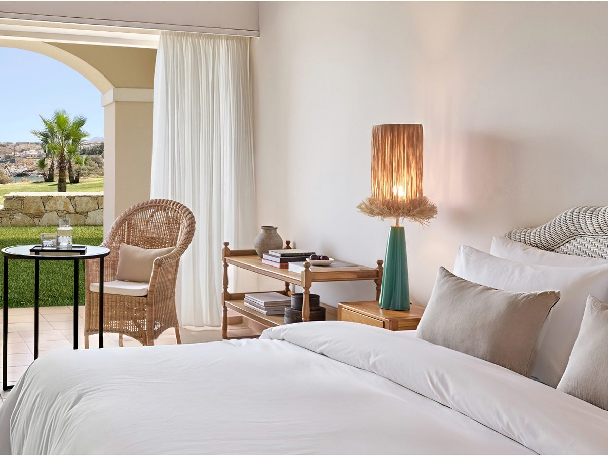 Bedroom with an arched opening to a lawn and palm garden, a wicker chair and a rattan bedside lamp.