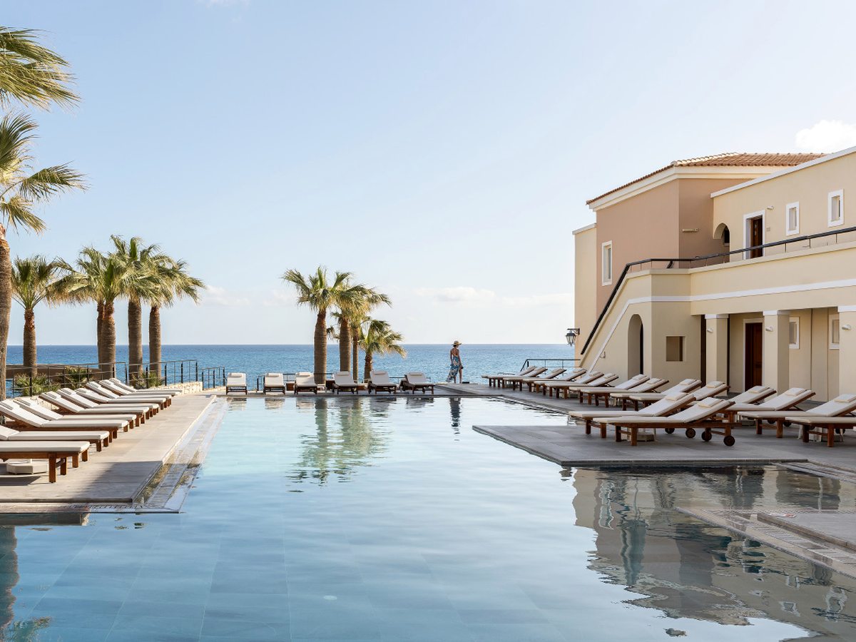 Rectangular pool at the seafront edge with sun loungers along one side, palms and cream resort buildings behind.