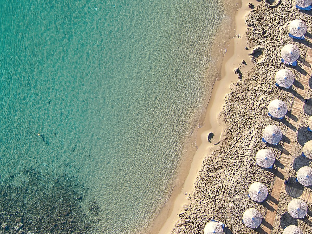 Overhead view of a sandy beach with rows of parasols and sunbeds beside clear turquoise shallows.