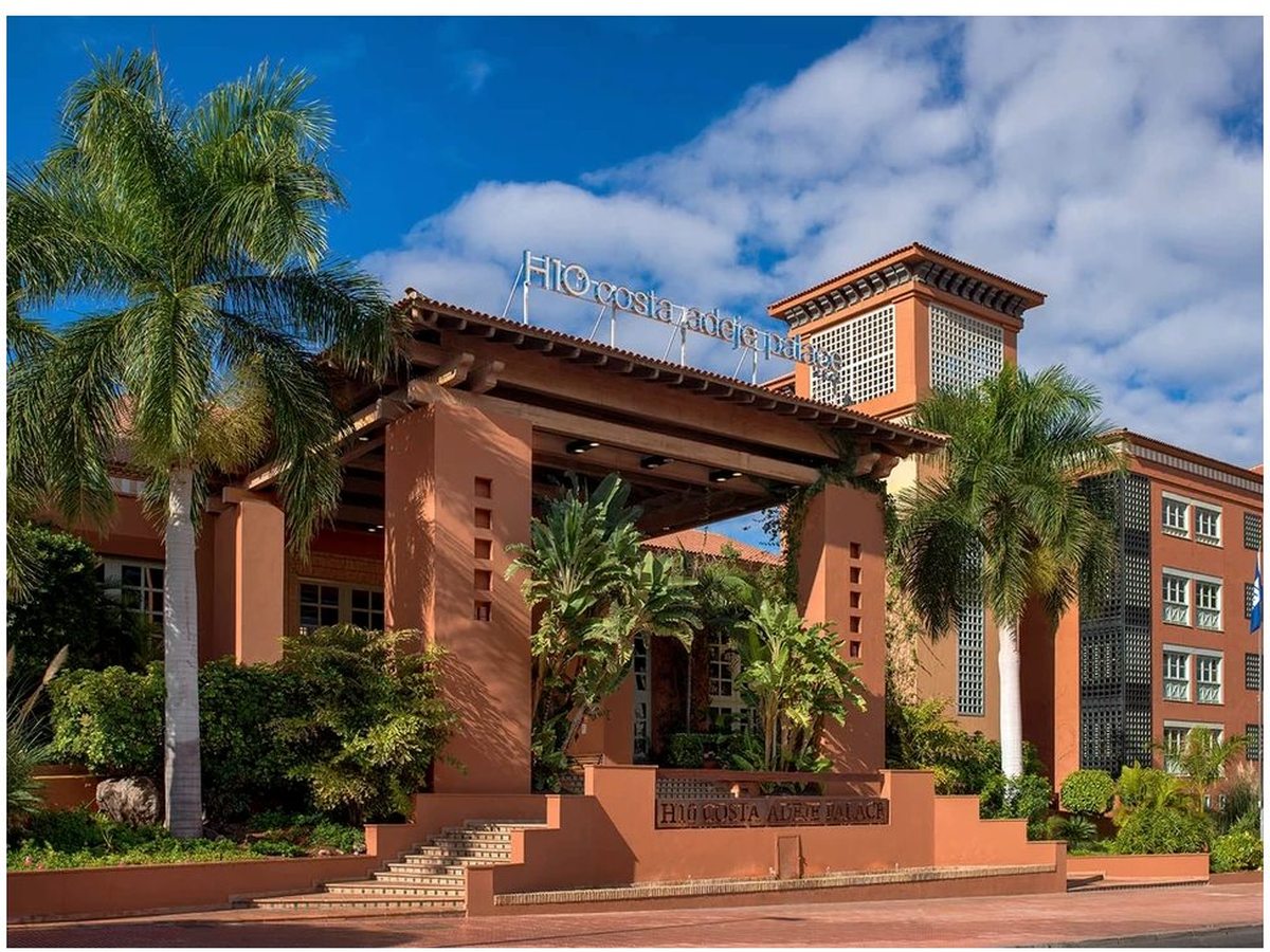 The terracotta entrance portico of H10 Costa Adeje Palace, framed by tall palms, the hotel's name mounted above.