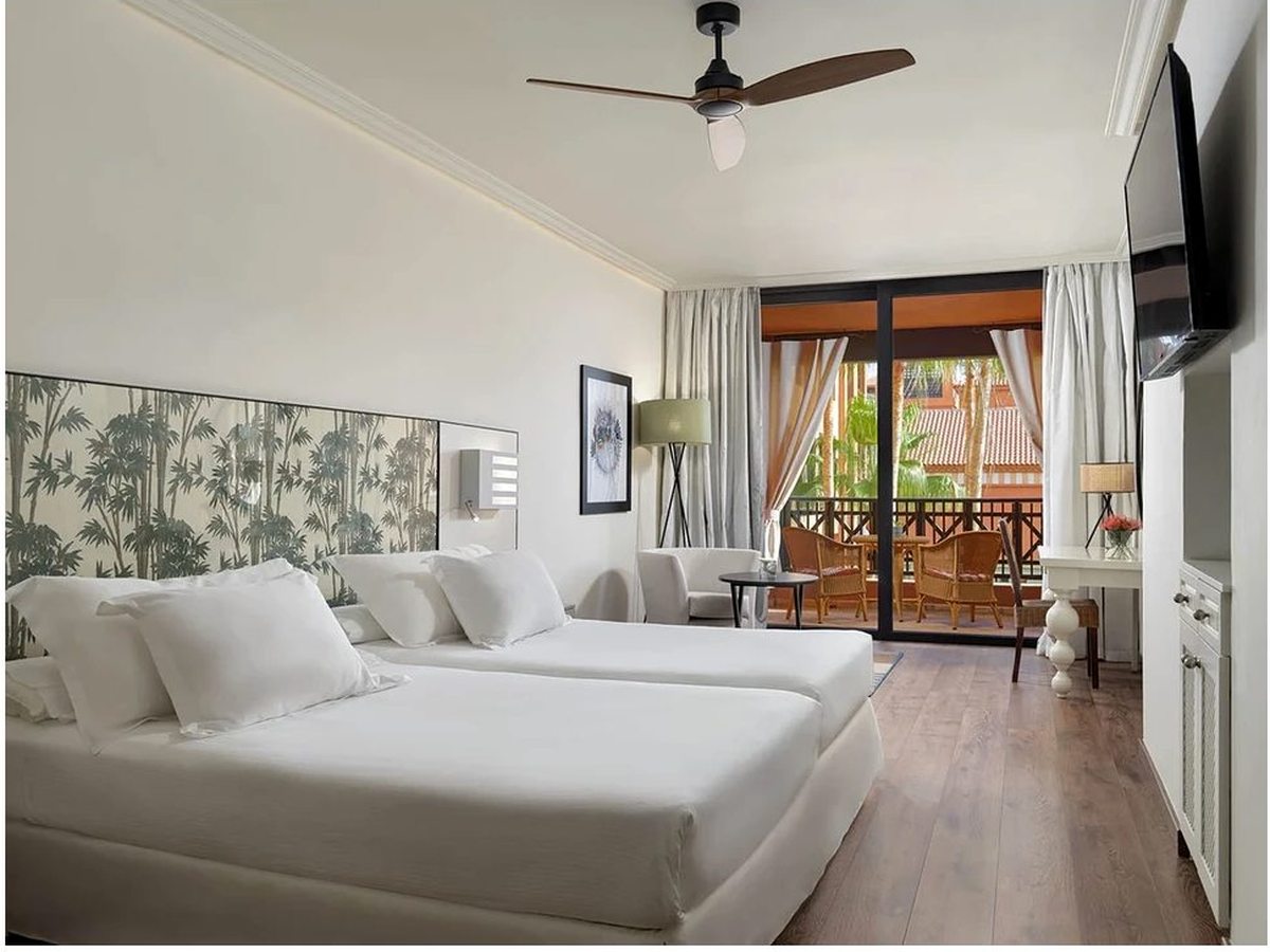 A double room with a patterned green headboard, a wall-mounted television and a curtained balcony beyond.