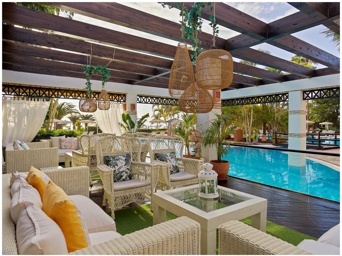 A covered poolside lounge with white wicker sofas and peacock chairs, cushions and hanging rattan lamps, beside the pool.