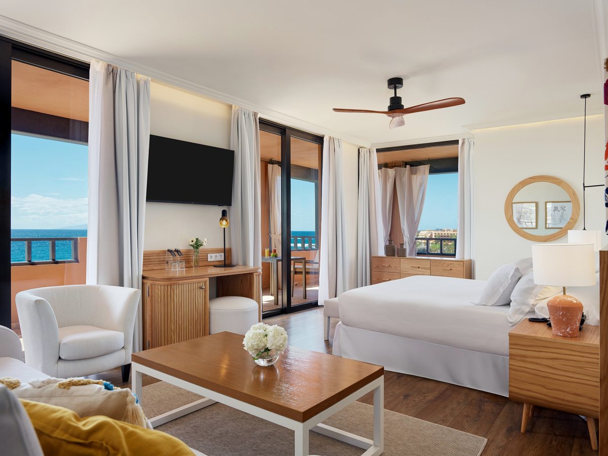 A guest room with a large bed, a small seating area and a balcony framing a side view of the sea.