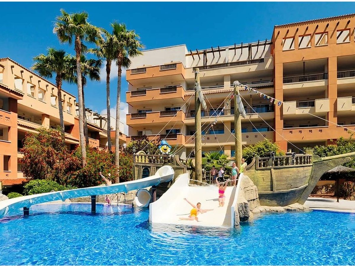 A children's pool with a wooden pirate ship and two white water slides, children sliding into the water, palm trees and terracotta apartment blocks behind.