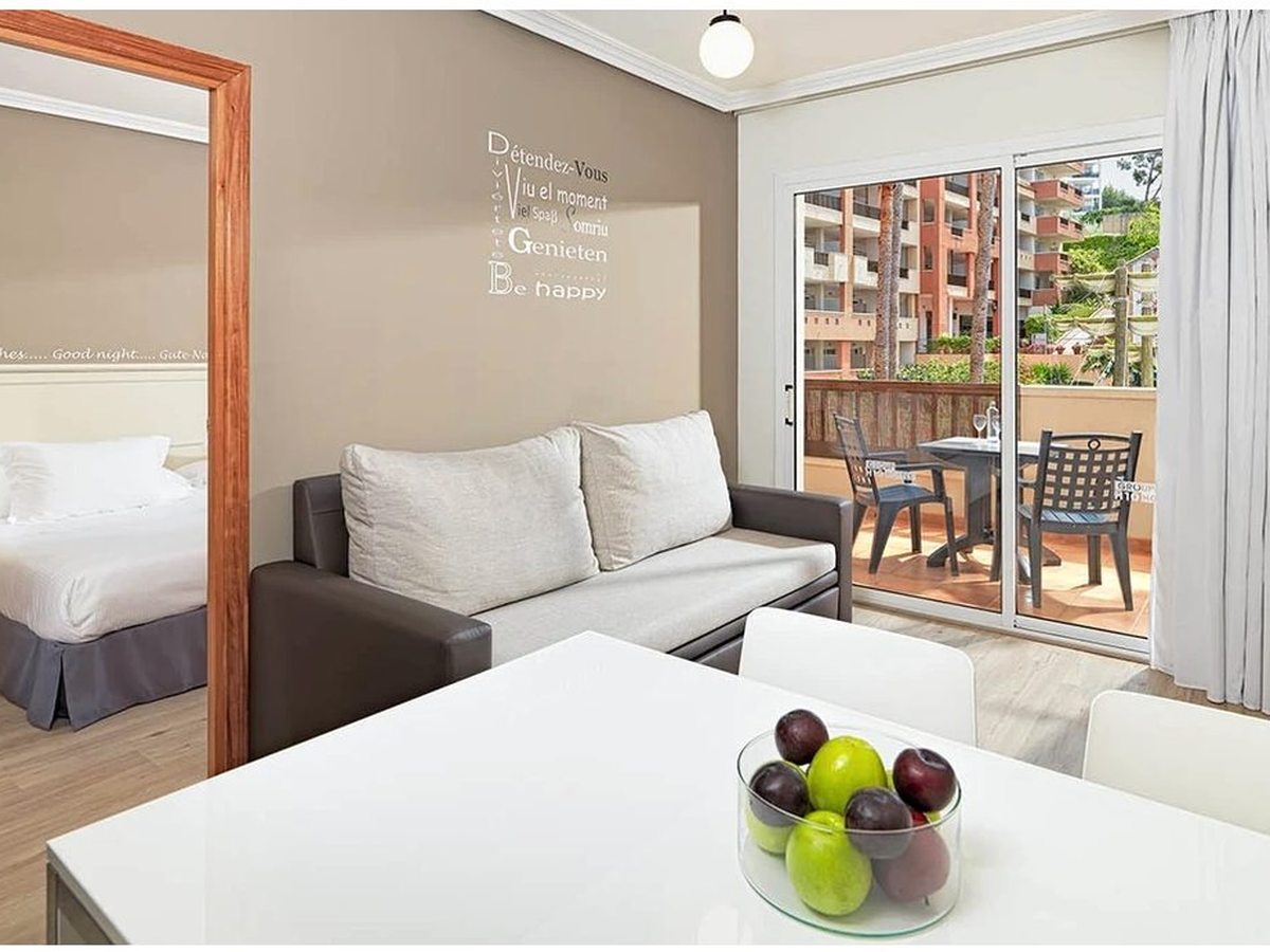 An apartment living room with a sofa and a white dining table, a bedroom visible through an open doorway, and sliding doors to a terrace with a table and chairs.