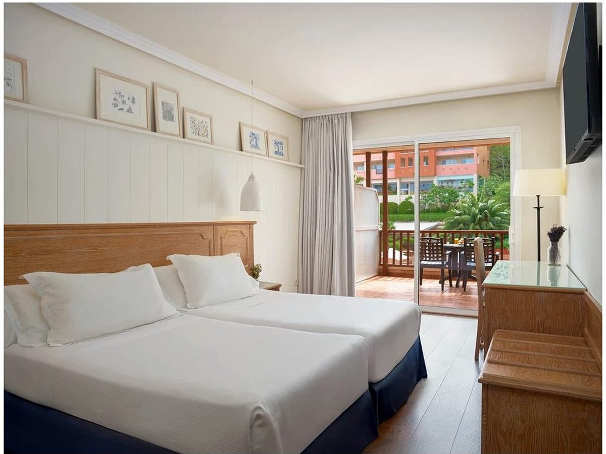 A hotel double room with a wooden headboard, framed pictures above the bed and sliding doors to a furnished balcony overlooking the resort gardens.