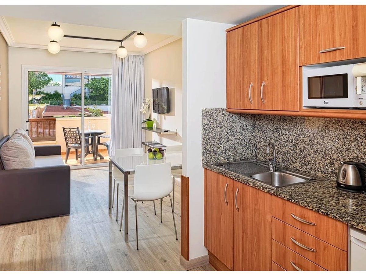 An apartment kitchen with light wood cabinets, a hob, microwave and sink along one wall, opening onto a living area with a sofa and dining table by the balcony.
