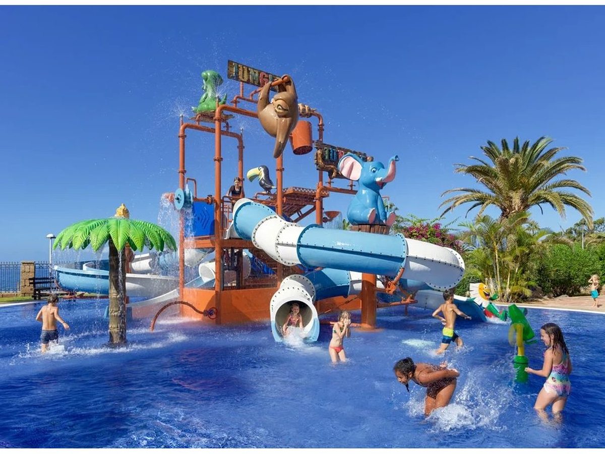 A jungle-themed splash park with a tall play tower, tipping bucket, blue tube slides and animal figures over a shallow pool where children play.
