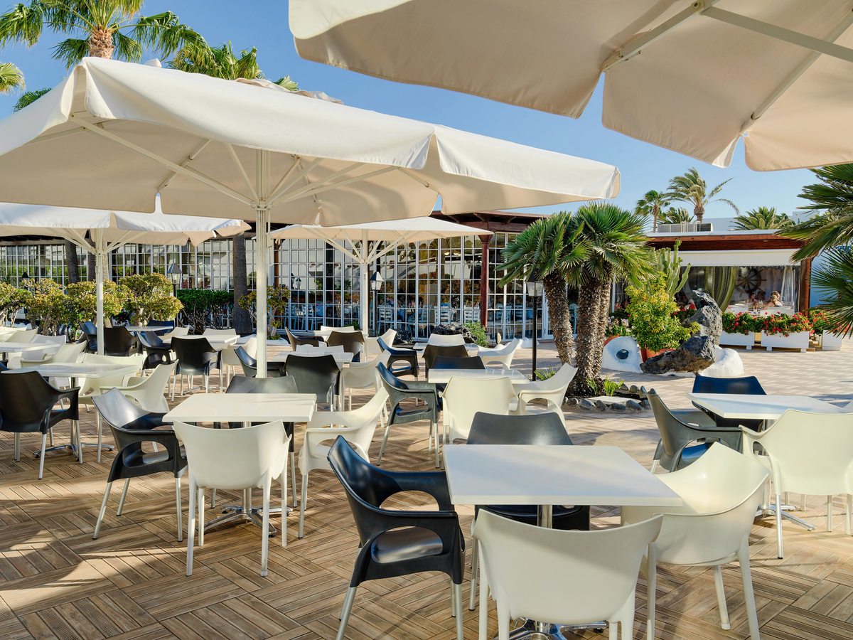 An outdoor terrace of white and black tables and chairs under large cream parasols, with palms and a glass-fronted building behind.