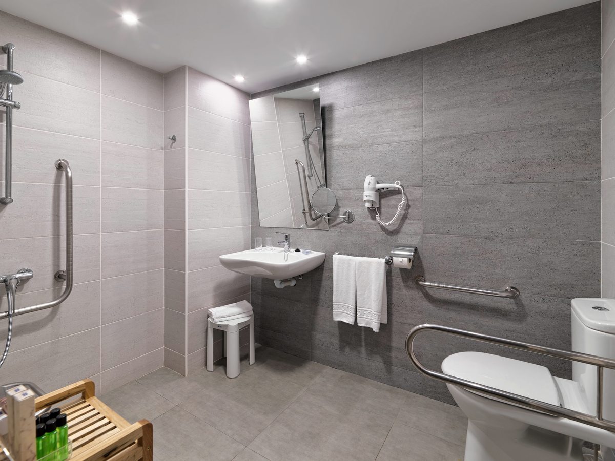 A grey-tiled bathroom with a level walk-in shower, grab rails beside the shower and the toilet, a wall-mounted basin and a wooden slatted bench holding toiletries.