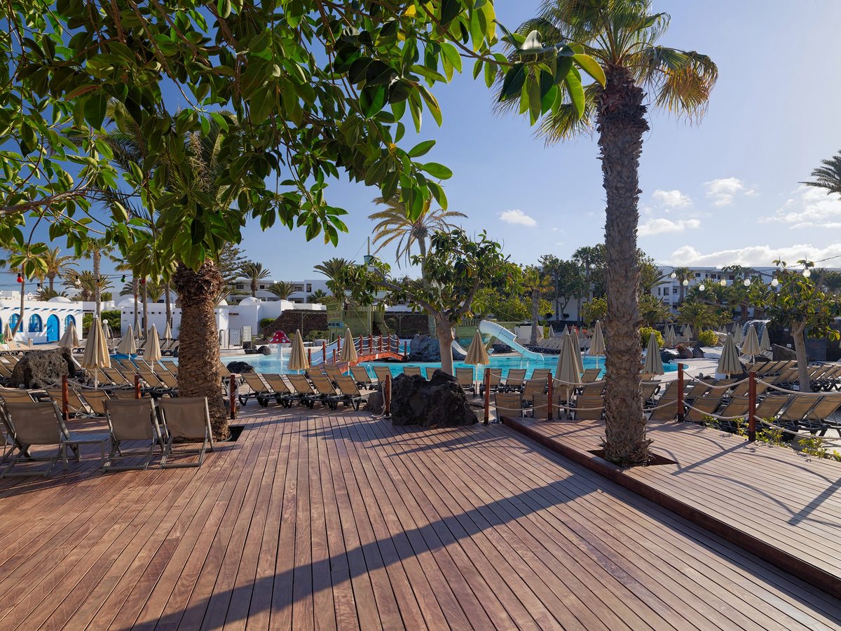A wide wooden sun deck with rows of loungers and folded parasols under mature palms, with a blue pool and a white slide behind.