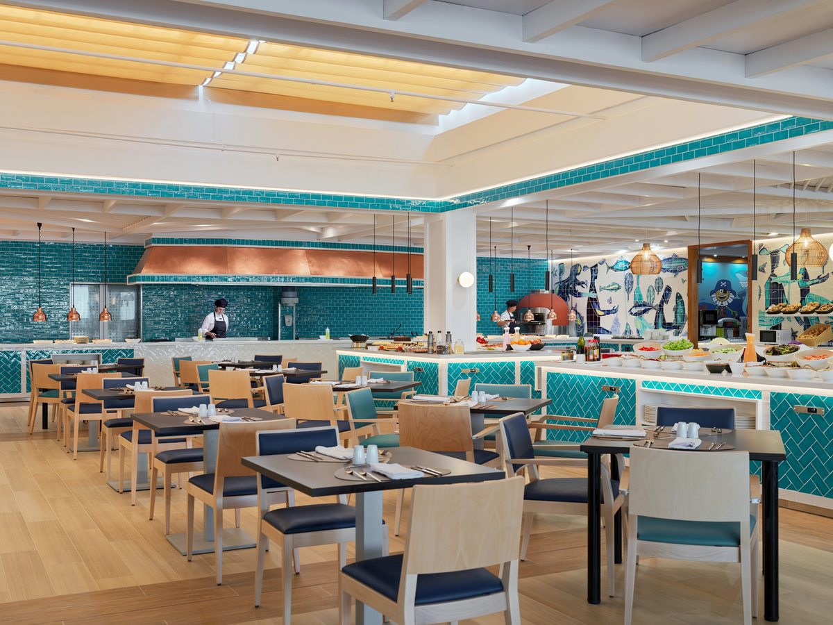 A bright buffet restaurant with pale wood tables and blue chairs, turquoise-tiled counters, a copper extractor hood over an open kitchen and a fish mural along the back wall.