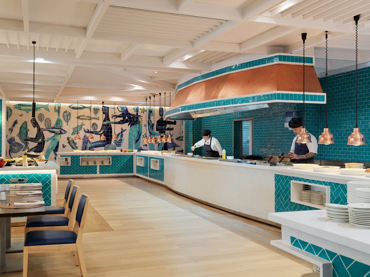 Two chefs working behind a white and turquoise-tiled counter under copper heat lamps, with stacked plates alongside and a blue fish mural on the wall.