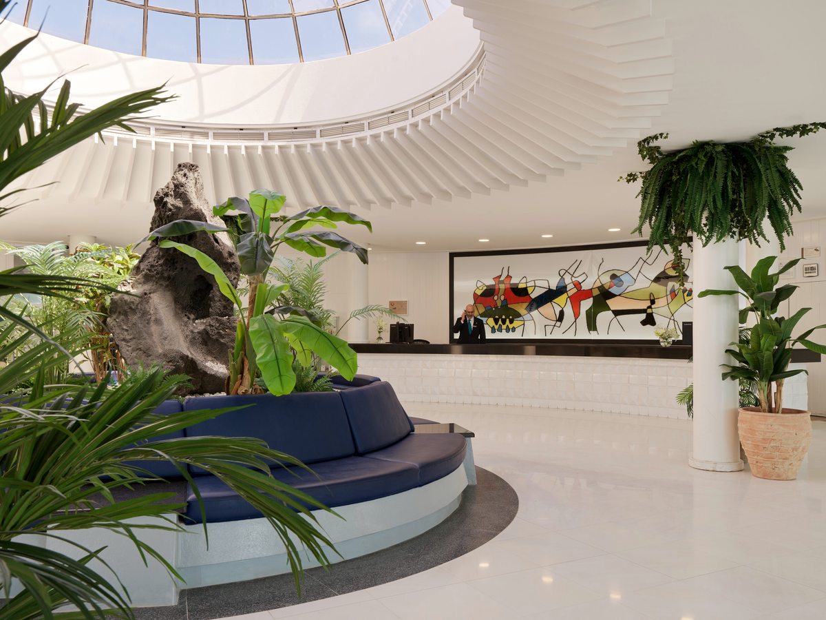 A white hotel lobby under a circular glass skylight, with a curved navy bench seat around a lava rock planter of tropical foliage and a long reception desk beyond.