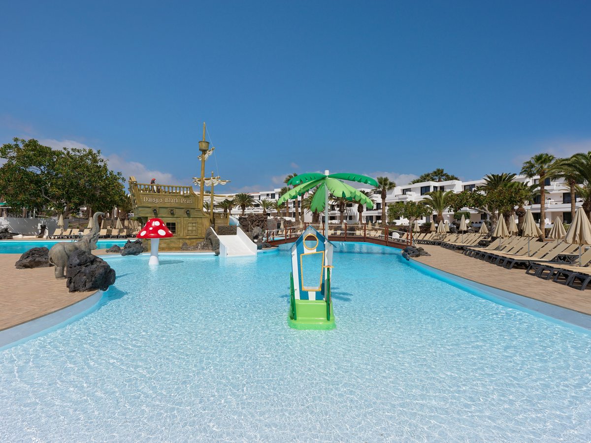 A shallow pool with a small play tower under a green palm-shaped parasol, a stone elephant sculpture at the water's edge and the pirate ship beyond.