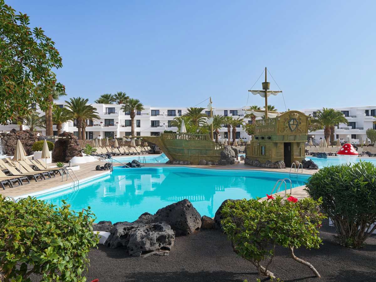 A large turquoise pool with a wooden pirate ship at its far edge, white low-rise resort blocks and palms behind, and volcanic rock and hibiscus in the foreground.