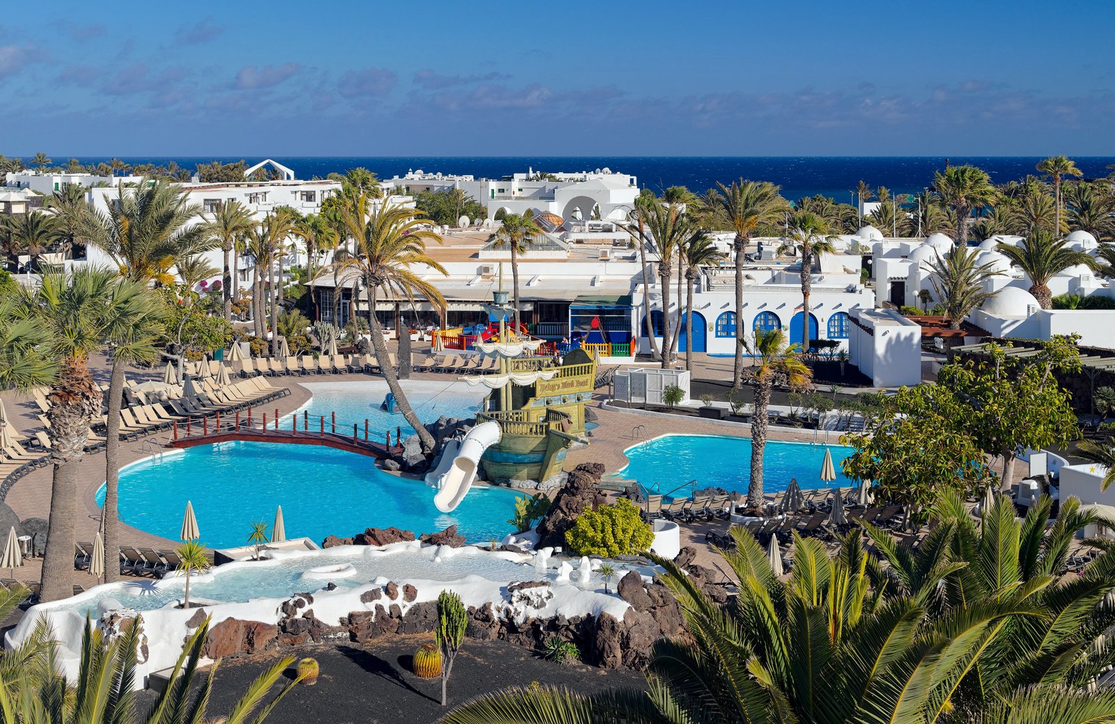 The pools at H10 Suites Lanzarote Gardens, with a wooden pirate ship and its white slide between them, white low-rise blocks and palms behind and the sea beyond.