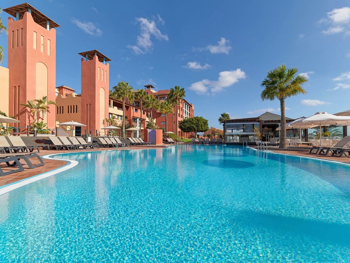A large curved outdoor pool edged with sun loungers and parasols, the resort's terracotta towers rising behind.
