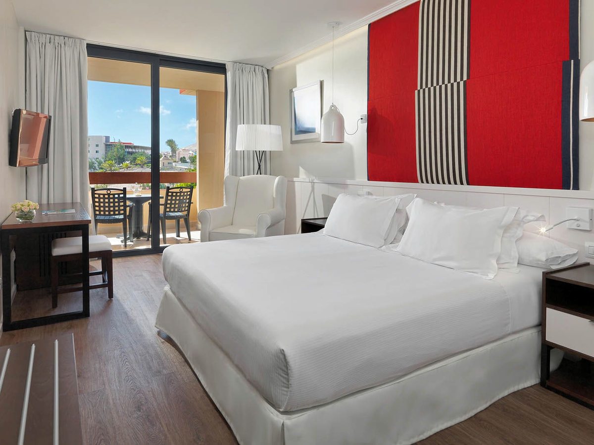 A room with a queen double bed, a red striped headboard panel, a desk and a balcony beyond.