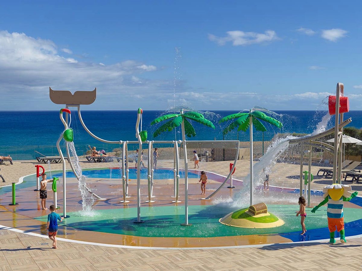 A circular splash park with palm-shaped sprays, a tipping bucket and children playing, the sea directly behind.
