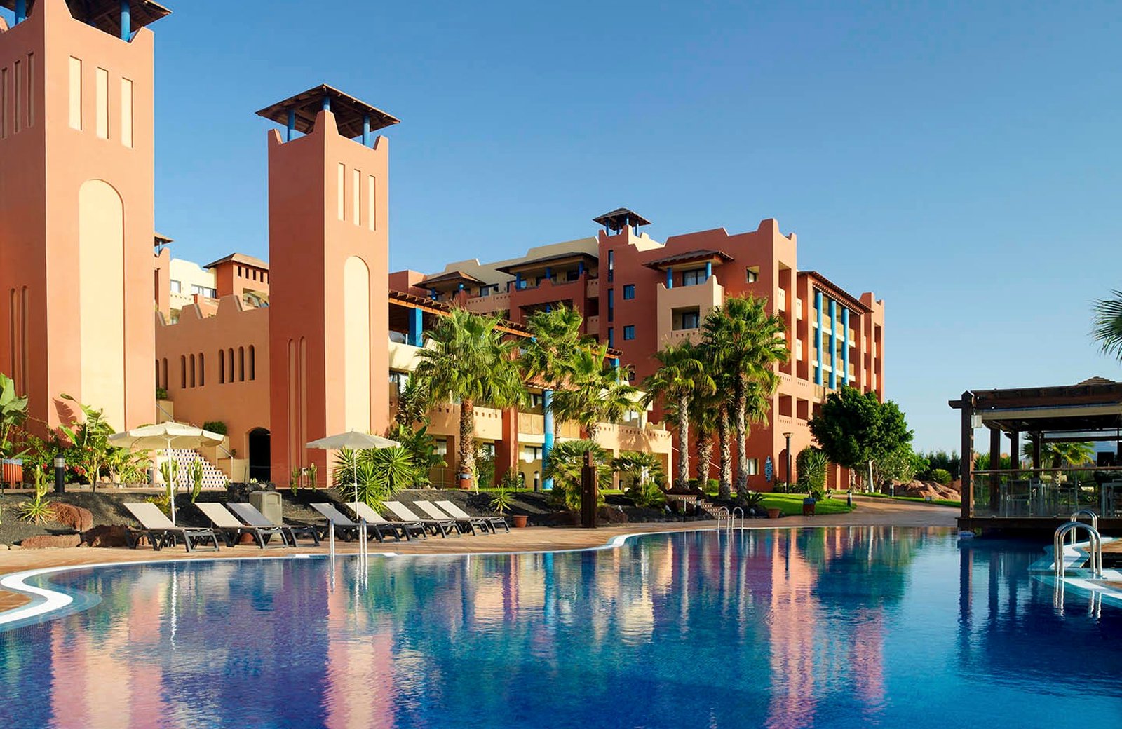 H10 Tindaya's terracotta-toned towers reflected in one of the resort's pools, framed by palms under a clear sky at Costa Calma, Fuerteventura.