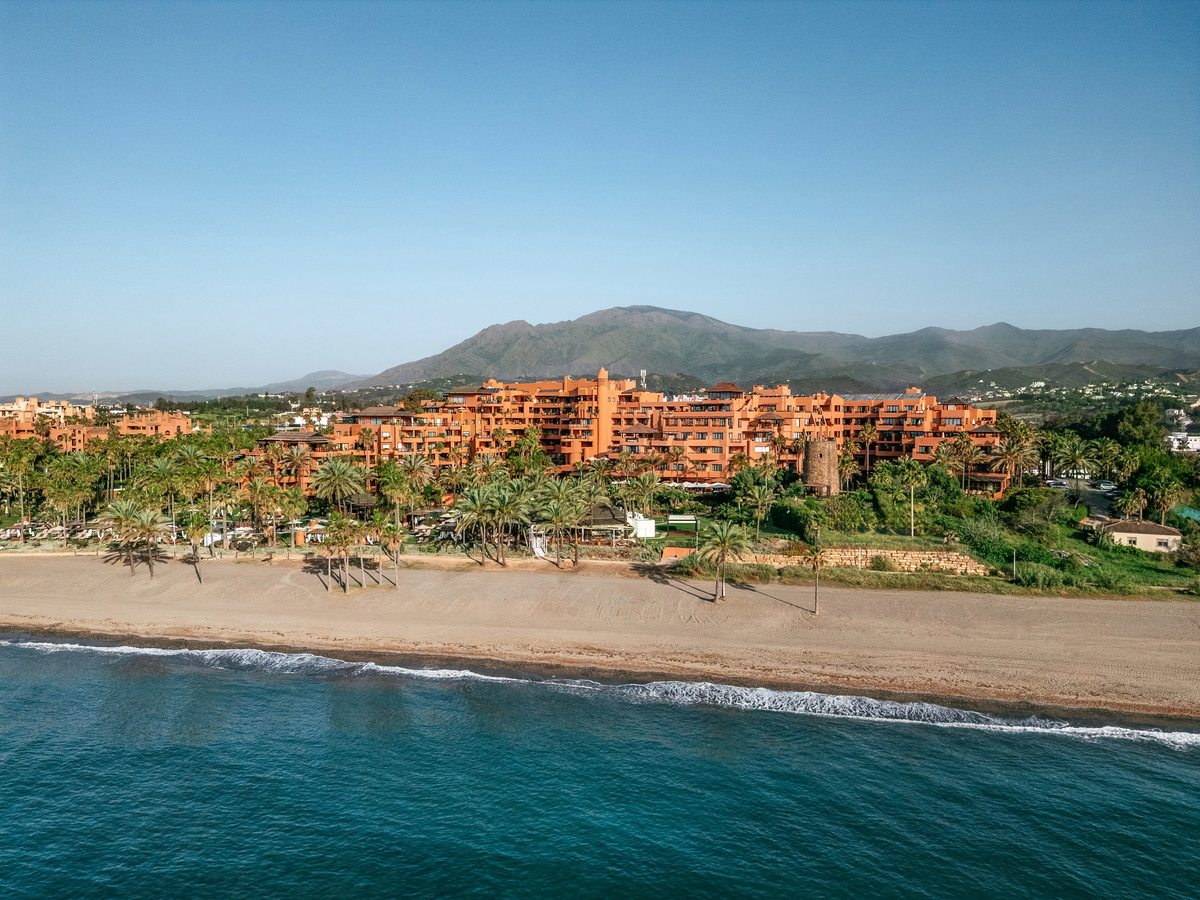 View along the shore of the terracotta resort buildings behind a palm-lined beach, with wooded hills rising behind.