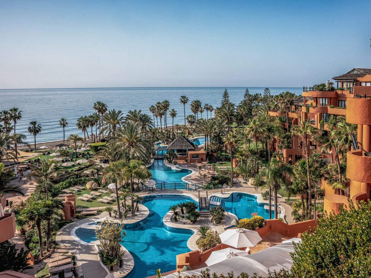 An elevated view over the resort's free-form pools set among lawns and tall palms, with the sea beyond.