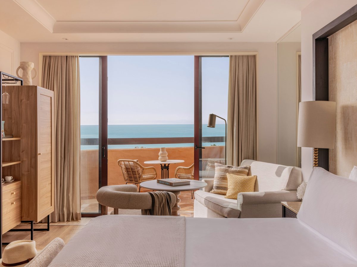 A bright room with a large bed and a sofa and armchair sitting area, glass doors opening to a balcony with a sea view.