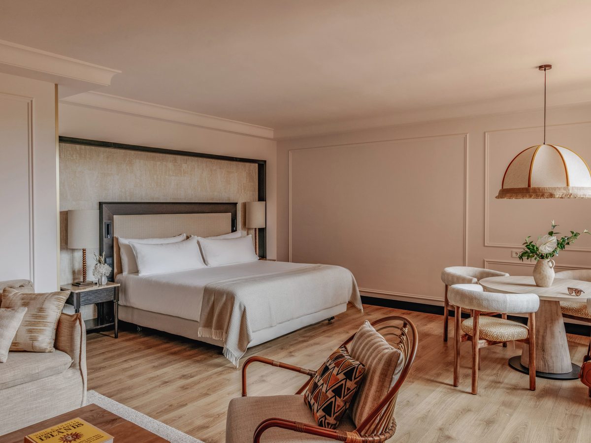 An open-plan suite with a bed, a lounge sofa and a round dining table in one large room, with a balcony beyond.
