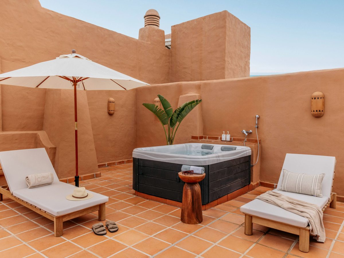 A walled terracotta-tiled terrace with an outdoor hot tub, two sun loungers and a parasol under a clear sky.
