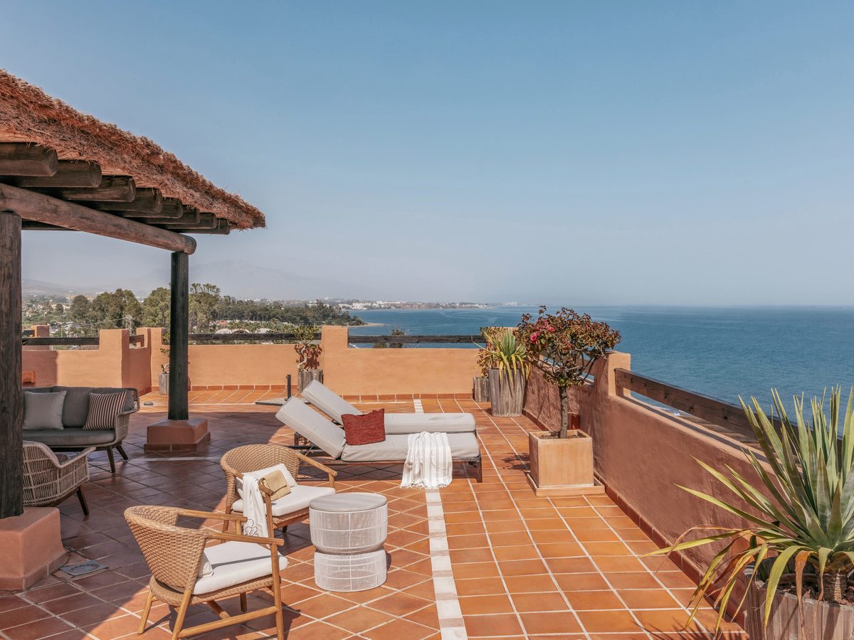 A terracotta rooftop terrace with a thatched pergola, sun loungers and wicker chairs looking along the coastline to the sea.