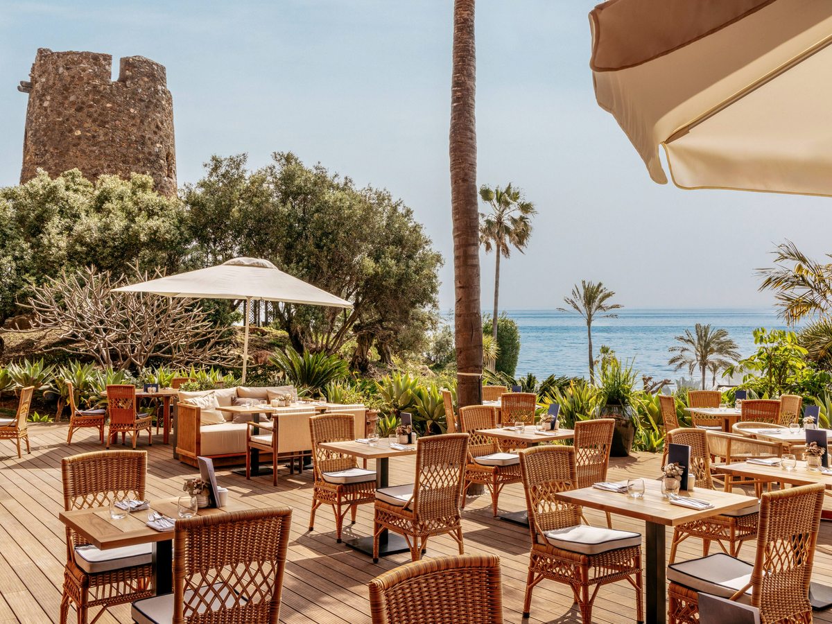 A decked garden terrace set with laid tables and parasols, palms and the sea beyond, with a stone watchtower to one side.