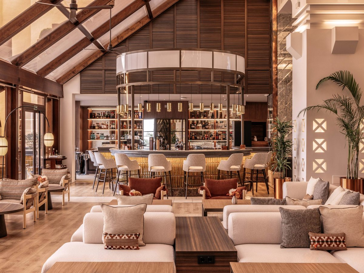 A double-height lobby bar with a long wooden counter, a bottle display, sofas and armchairs under a raftered ceiling.