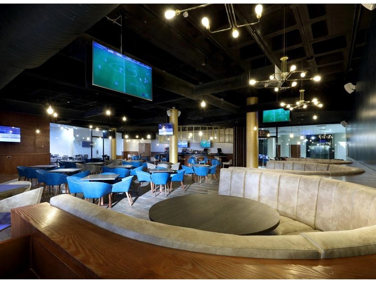Sports bar with wall-mounted screens showing football, blue chairs at high tables and a curved central bar.
