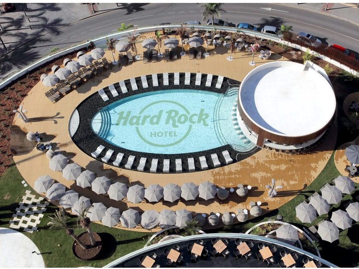 Aerial view of an oval pool tiled with the Hard Rock Hotel logo, ringed by sun loungers and parasols.