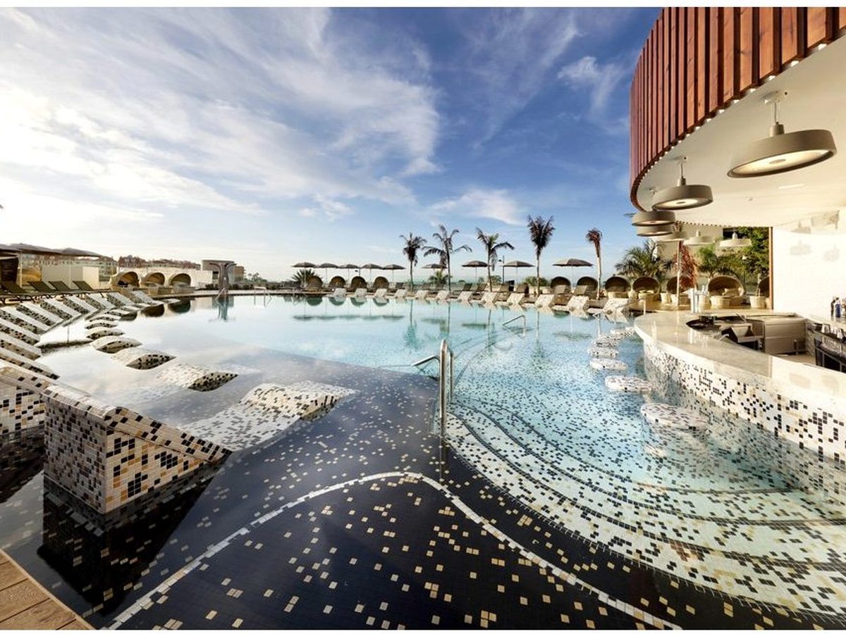 An infinity pool with submerged loungers and mosaic tiling, its edge open to the sea, palms and parasols around the deck.